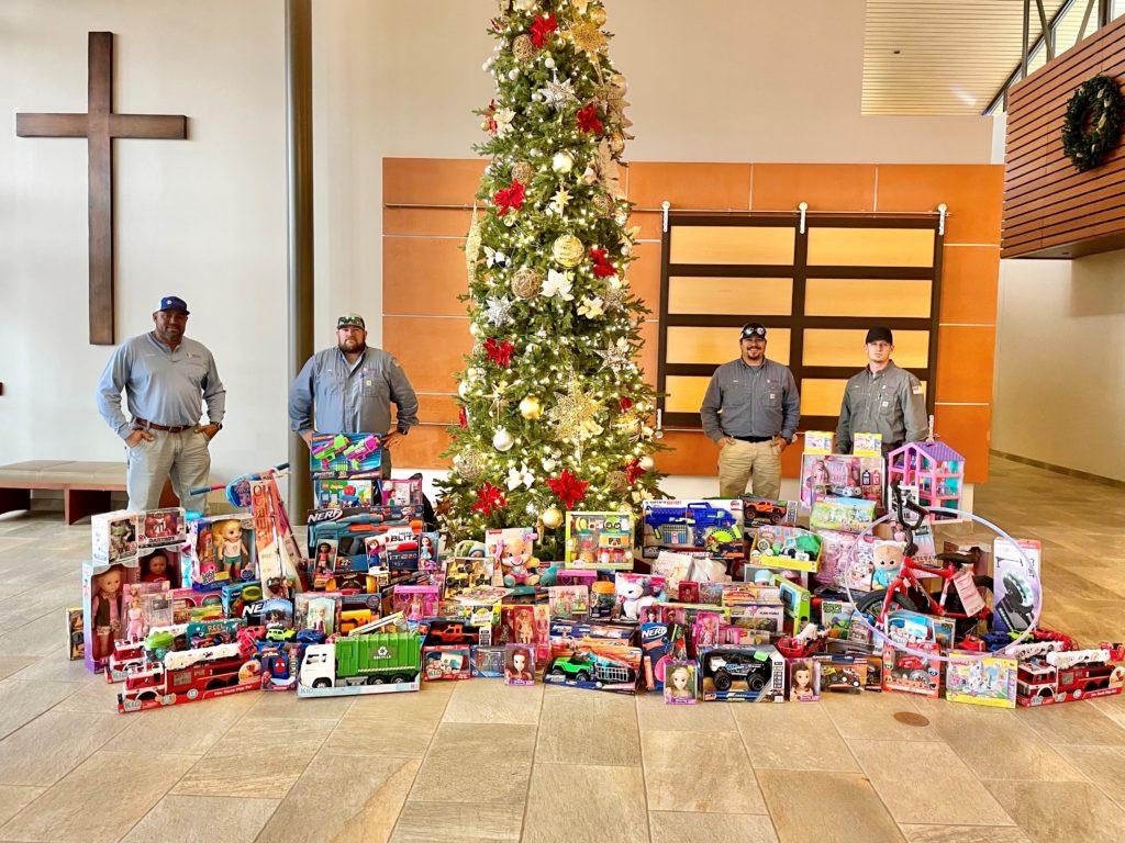 Four men in front of a Christmas tree with lots of presents they donated.